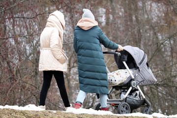 Fototapeta na wymiar Two young women with a baby stroller walking in a park. Early spring weather, concept of motherhood, couple of girlfriends with pram