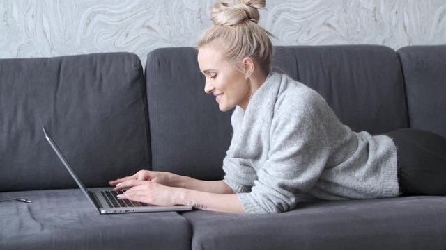 Happy blond woman lying prone on sofa and working on laptop computer