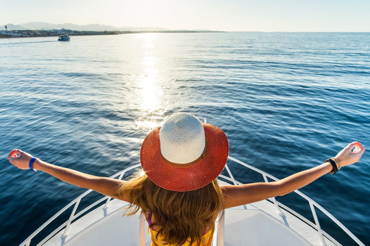 Young woman with long hair wearing yellow dress and straw hat standing with raised hands on white yacht deck enjoying view of blue sea water.