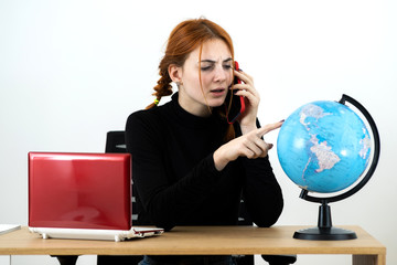 Young travel agent woman sitting behind working desk with laptop computer and geographic globe of the world talking on a cell phone.