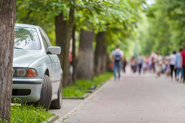 Car parked in pedestrian zone under trees along street with walking people on summer day.
