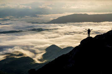 Wide mountain panorama. Small silhouette of tourist with backpack on rocky mountain slope with raised hands over valley covered with white puffy clouds. Beauty of nature, tourism and traveling concept