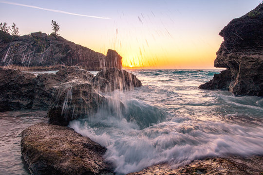 Water droplets spray from a large wave crashing on the rocky coast. Sun just above the horizon in the background - Jobsons Cover Bermuda