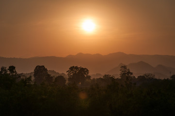 silhouettes of trees and layered mountains in an orange light with a setting sun in the background