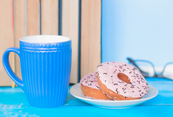 background with books, a Cup of coffee and doughnuts. a Desk with a blue coffee Cup, a stack of books, glasses, and doughnuts.