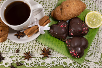 On a white saucer is a cup of coffee, cinnamon sticks. On a green saucer lies cookies, croissant and lemon.