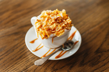Hot caramel latte with cornflakes over milk foam in coffee glass on white plate with sweet caramel art on wooden table.