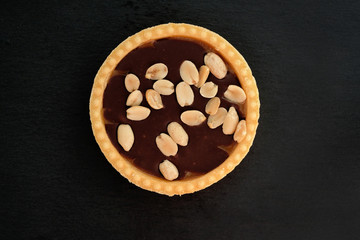 Sweet tartlet with chocolate and peanuts on top. Shortcrust pastry cake with nuts and chocolate fondant on a black background.