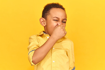Isolated shot of emotional disgusted African boy smelling something stinky and disgusting, pinching nose. Displeased child can't stand intolerable smell, holding breath with fingers on his nose - obrazy, fototapety, plakaty