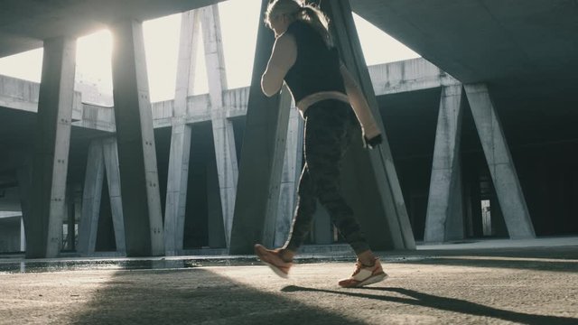 Athletic woman doing walking lunges outside in abandoned urban garage building. Side tracking shot in slow motion with sunlight in background.