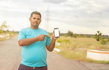 Young man wearing jogging dress showing smartphone screen by pointing with hand and finger at park - concept of recommended fitness tracker app on mobile.