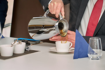 Hand of a businessman in a suit and tie, sitting at the table and pouring tea from a silver thermos...