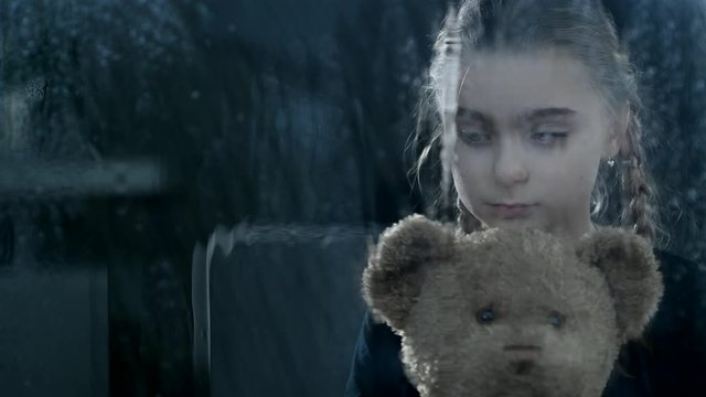 Worried Girl Cuddles Closely With Her Teddy Bear. The Storm Makes The Waiting Room Look Dark And Gray.