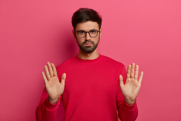 Photo of European man with thick stubble makes stop gesture, hold sign, pulls palms towards camera, looks seriously, has determined expression, asks to calm down, wears red sweater, glasses.