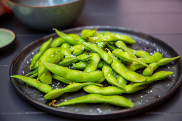 Cooked green Edamame lie on a dark plate in a Japanese restaurant.