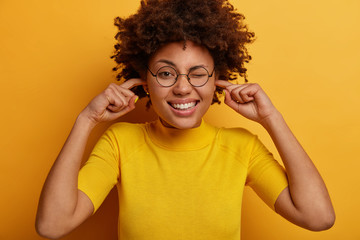 Funny dark skinned woman plugs fingers in ears, cannot concentrate because of noise in crowded place, winks eye and shows white teeth, wears spectacles and tshirt, isolated on yellow background