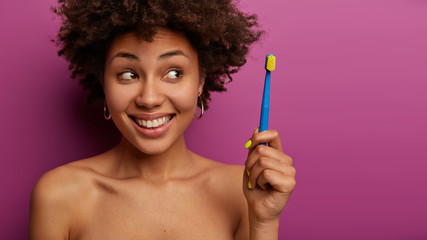 Positive curly haired woman looks happily aside, has Afro hairstyle holds toothbrush, happy mood, stands naked against purple background, cares about teeth, cleans regularly, smiles broadly. Hygiene