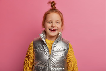 Positive redhead girl laughs happily, wears warm knitted sweater and vest, enjoys spending free time with parents, has happy gaze at camera, feels carefree, isolated over rosy pastel background