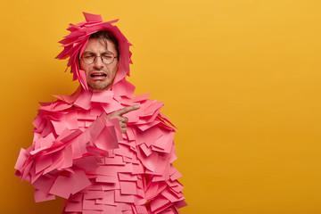 Upset dejected man cries desperately, points away on blank space, dissatisfied with sales discounts, many pink stickers stuck over body, isolated on yellow background. Unhappy desperate feelings