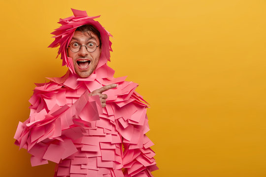 Positive funny man indicates on blank space at upper right corner, advertises some product, wears outfit made of adhesive notes, round spectacles, isolated over yellow background. Look at this