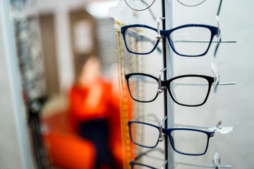 Row of glasses at an opticians. Eyeglasses shop. Stand with glasses in the store of optics. Woman chooses spectacles. Eyesight correction.