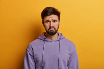 Fototapeta premium Unamused sad miserable guy purses lips, frowns face with dissatisfaction, wears hoodie, poses indoor against yellow background, sulks troubled as someone hurt his feelings, wears purple hoodie