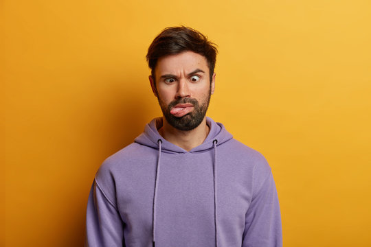 Serious crazy bearded man crosses eyes and sticks out tongue, has funny grimace, feels bored, raises eyebrows, wears casual hoodie, isolated on yellow background. Human face expressions concept
