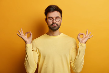 Photo of relaxed unshaven European man stands in lotus pose, makes zen gesture, breathes deeply and...