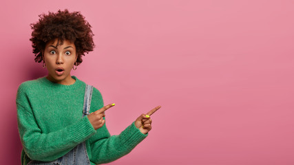Photo of surprised curly haired young woman indicates away, shows free space for your promotion, fascinated by high prices, gasps from wonder, wears green sweater, isolated on pink background - obrazy, fototapety, plakaty