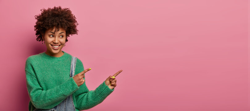 Positive ethnic woman points at product, suggests check it out, indicates right with happy smile, rejoices nice advertisement, wears green sweater, poses against pink background. Promo concept