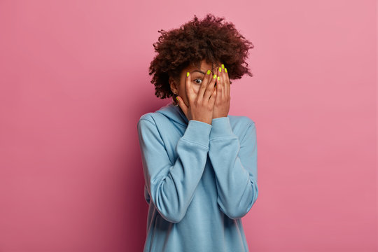 Frightened African American woman covers face with palms, stares through fingers, being afraid of something, wears blue sweatshirt, hides herself from something terrifying, isolated on pink wall