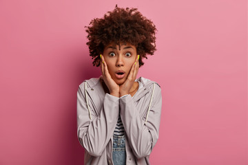 Fototapeta na wymiar Horizontal shot of curly woman stares with shock and embarrassement, shocked to get surprising news, touches cheeks, gasps with wonder, wears casual anorak, poses against pink studio background