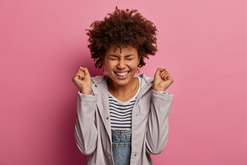 Joyful emotive ethnic curly woman clenches fists, celebates victory, smiles broadly, being in high spirit, closes eyes, wears casual anorak, rejoices achieve goal, poses against pink background.