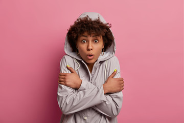 Horizontal shot of shocked young Afro American woman crosses arms, wears anorak with hoody, feels frightened, cold and trembles from fear, afraids of something, poses over pink studio background