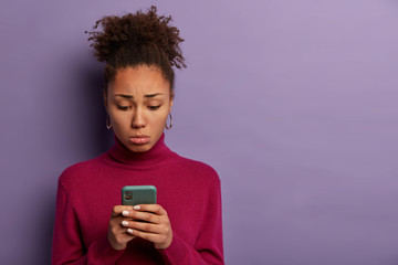 People, unhappy emotions concept. Sad disappointed woman received bad sms, holds smartphone and concentrated at screen with dissatisfaction, wears burgundy poloneck, poses indoor, feels lonely