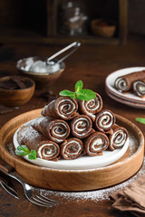 Rolled chocolate pancakes with ricotta filling on a white plate on a wooden table. Chocolate crepes.  Selective focus.