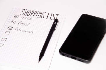 Shopping list. . Squared notebook with black pen on a white background. Record ideas, notes, plans, tasks. The list includes bread, milk, bananas.  Smartphone with a calculator.