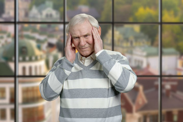 Old caucasian man massaging temples. Grandfather with terrible headache in windows background with view on the town.