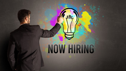 businessman drawing colorful light bulb with NOW HIRING inscription on textured concrete wall, new business idea concept