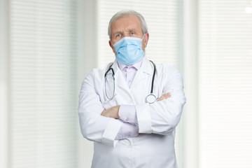 Serious doctor with medical mask and crossed arms. Portrait of senior experienced physician in...
