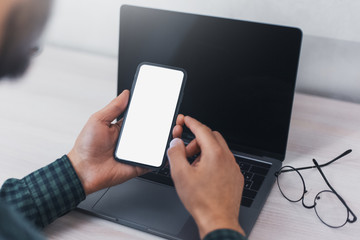 Close-up of male hands, holding smartphone on background of laptop on desk, with mockup.