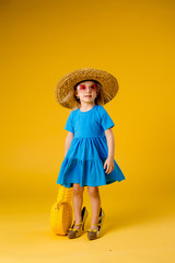 little blonde girl in a straw hat and sunglasses is smiling on a yellow background. child girl in a blue dress is holding a pineapple-shaped drink glass on a yellow background.summer vacation concept