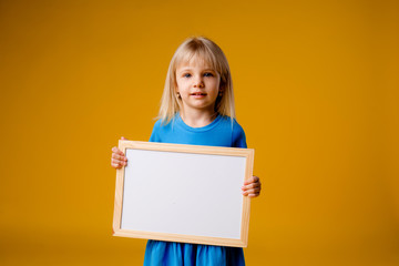 little blonde girl in a blue dress and a striped hat holds a white record board on a yellow background