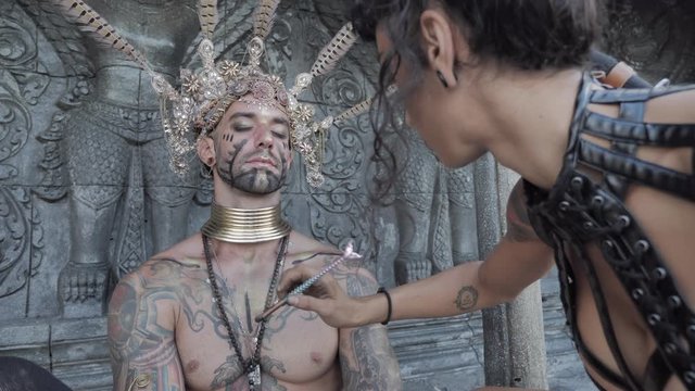  Girl in a tomb raider style prepares a man make up for a ethnic priest performance