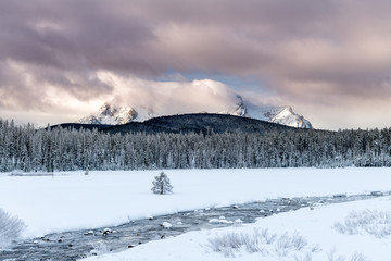 Fringed cold river flows across a snow covered meadow with high mountain peaks