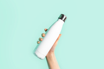 Close-up of female hand, holding white reusable steel stainless eco thermo water bottle, isolated on background of cyan, aqua menthe color. Be plastic free. Zero waste.