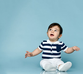 Little child in striped t-shirt, white pants and booties. He is looking up, sitting on floor against blue background. Close up