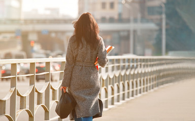 Pretty woman in coat with books in hand walking on the bridge. Young stylish student walks on the bridge. Female with books hurries to classes, exam.