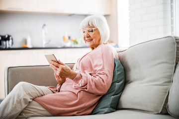 Cheerful old lady sitting on couch and using cellphone