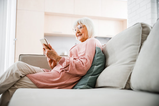 Joyful senior lady sitting on couch and using mobile phone